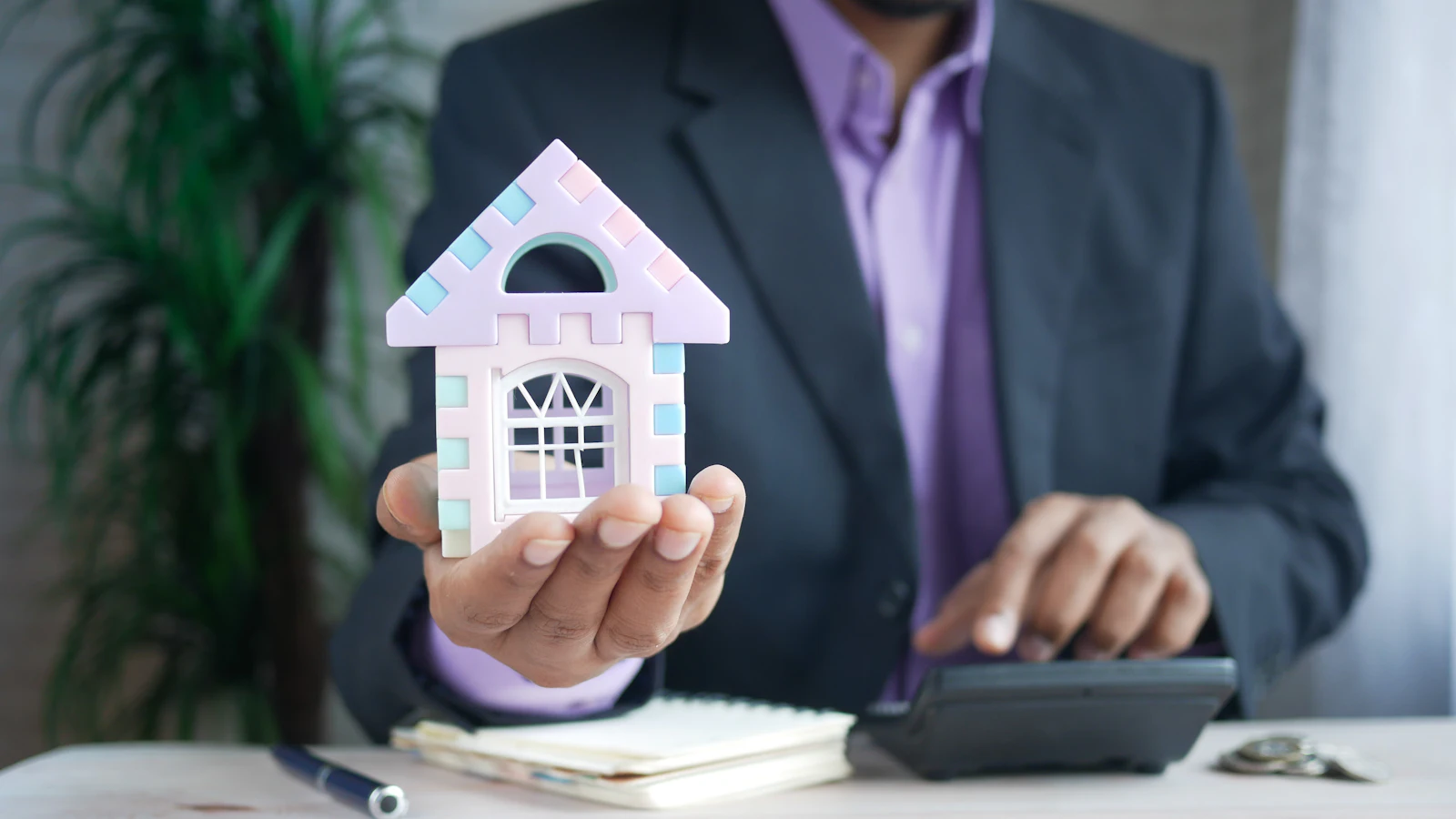 A person at a desk holding a model house next to a calculator and paperwork