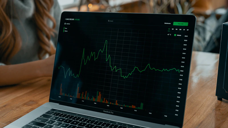 A laptop on a wooden desk displaying a stock price line chart with volume bars