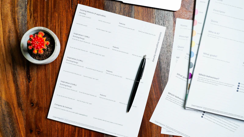 An overhead view of printed enrollment forms and a pen laid out on a plain wooden desk, with the edge of an open laptop visible