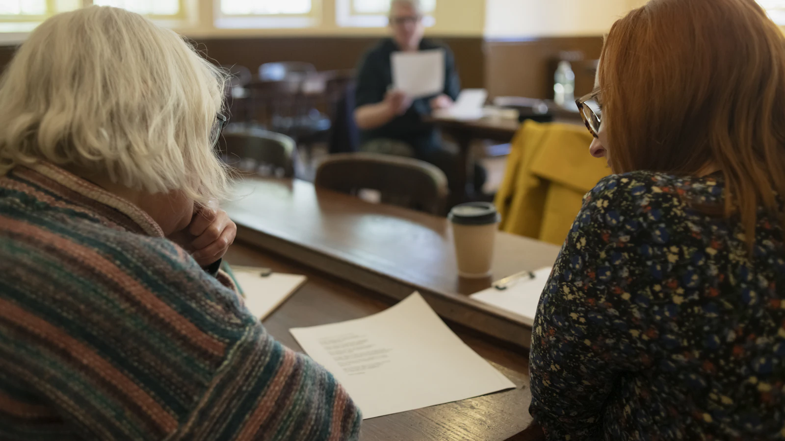An older woman and a younger woman sitting close together at a table, both looking down at a printed document, with a coffee cup nearby