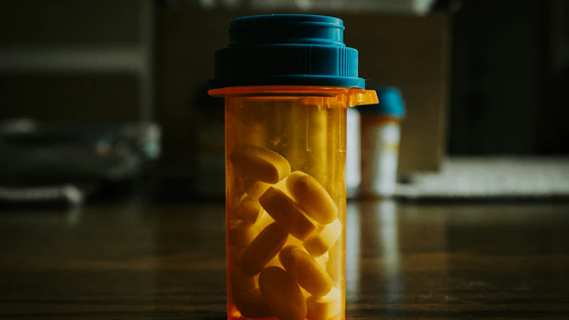 An open amber prescription pill bottle filled with tablets, sitting on a dark wooden desk